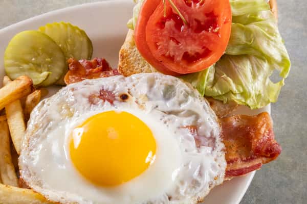 Breakfast sandwich with sunny-side-up egg, bacon, lettuce, and tomato on toasted bread, served with fries and pickle slices.
