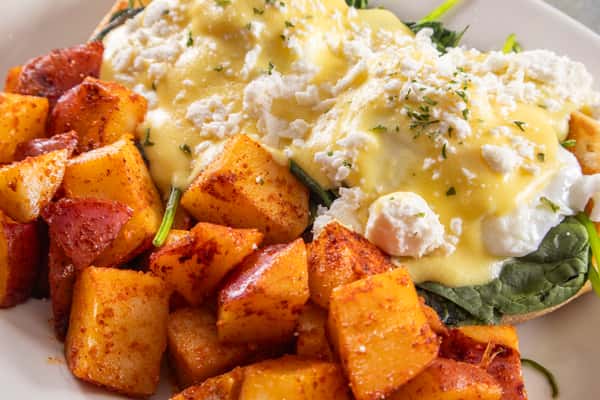 Eggs Benedict with poached eggs, hollandaise sauce, and feta over spinach on English muffins, served with seasoned home fries.