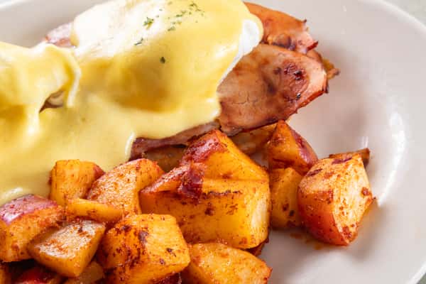 This image features a classic Eggs Benedict dish served with a side of seasoned breakfast potatoes.