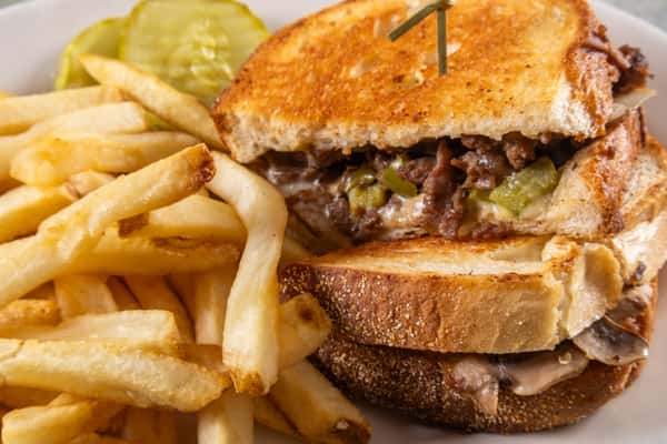 Grilled sandwich filled with beef, green peppers, and melted cheese, served with crispy fries and pickle slices on the side.