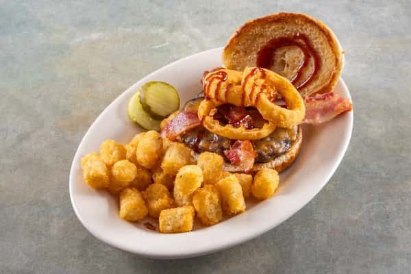 This dish is a loaded barbecue bacon cheeseburger topped with crispy onion rings and barbecue sauce, served with a side of golden tater tots and pickle slices. A hearty, classic diner meal!