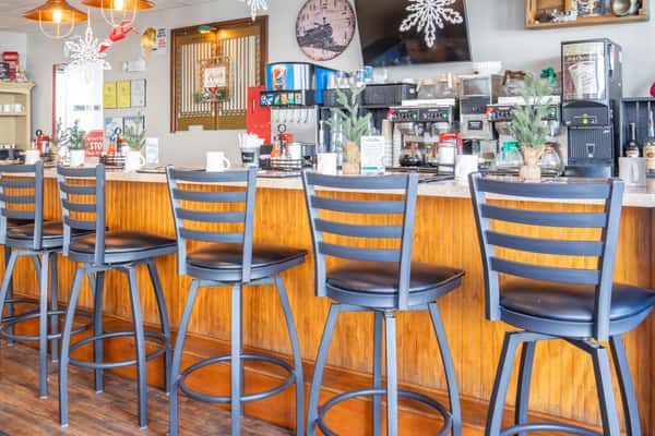 Five black bar stools lined up at a wooden counter with coffee machines, soda fountain, and holiday decor in a cozy diner setting.