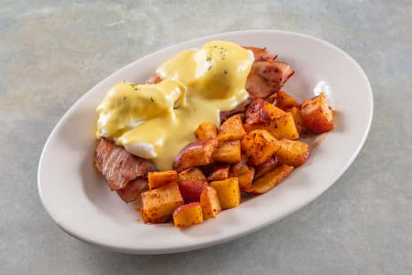 Classic eggs Benedict with hollandaise sauce, served over ham and paired with seasoned breakfast potatoes on a white plate.