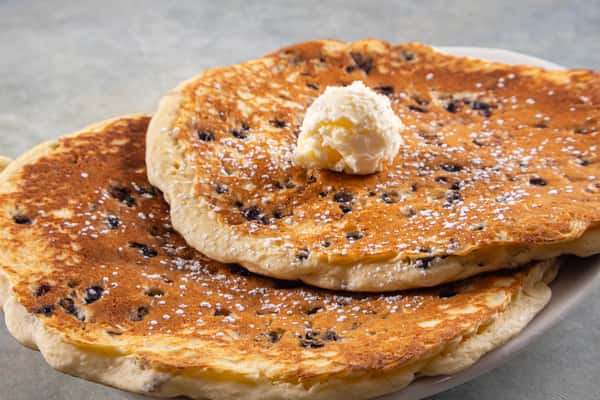 This dish features a stack of two large chocolate chip pancakes topped with a generous scoop of butter and a light dusting of powdered sugar. A sweet and indulgent breakfast choice!
