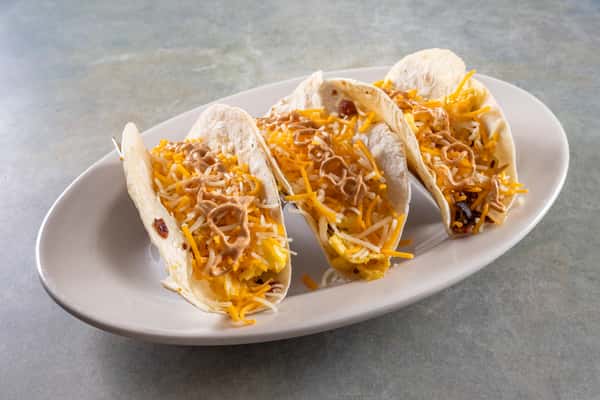 Four breakfast tacos with scrambled eggs, shredded cheese, and sauce in soft flour tortillas on a white oval plate.