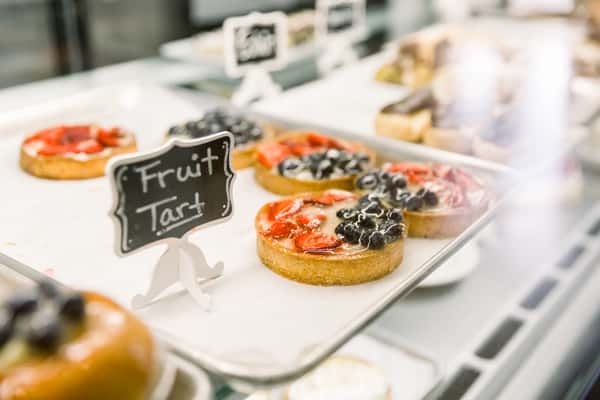 Display case with individual fruit tarts topped with strawberries and blueberries, labeled with a small "Fruit Tart" sign in a bakery setting