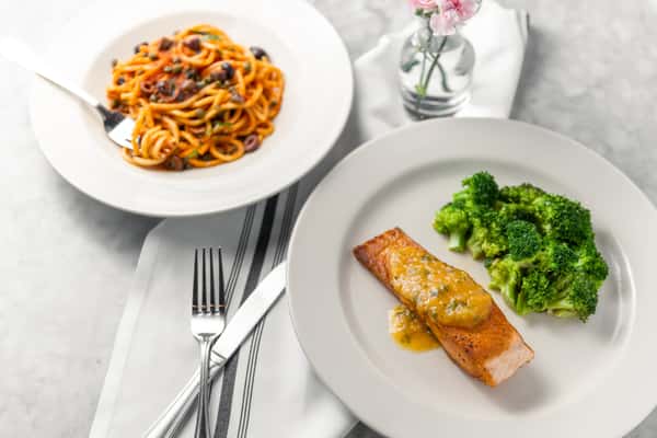 Top-down view of a plated meal featuring a grilled salmon fillet topped with a light sauce, served with steamed broccoli. In the background is a bowl of pasta with tomato sauce, olives, and capers, along with utensils and a small vase of pink flowers on a marble table.