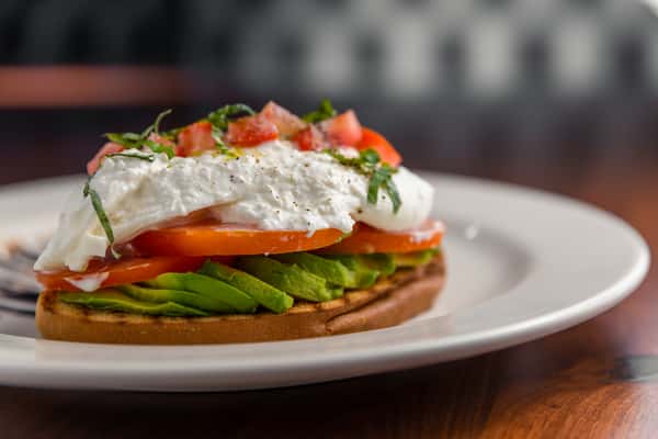 Avocado toast topped with sliced tomato, fresh burrata, and basil on a white plate.