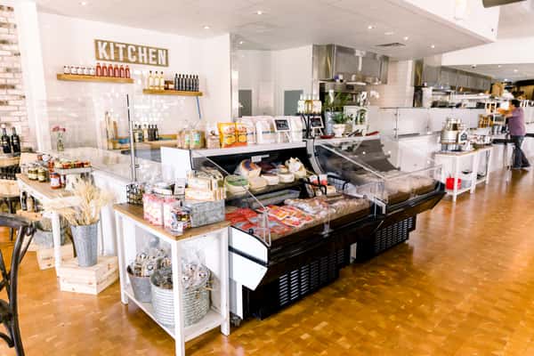 Bright, modern market-style café interior with deli display cases, open kitchen, wooden floors, and shelves stocked with specialty foods and wine