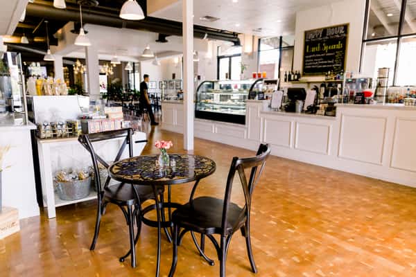 Bright, airy café interior with white counters, glass display case, chalkboard menu, and small round table with two chairs in the foreground