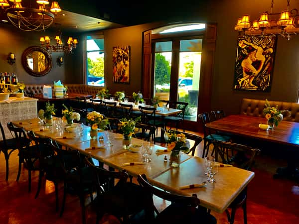 Warm, intimate restaurant dining room with set tables, floral centerpieces, chandeliers, and large windows letting in natural light