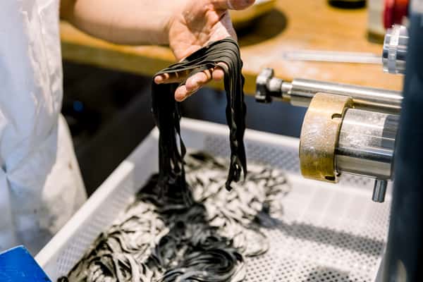 Fresh black pasta noodles being extruded and cut from a pasta machine, held in a cook's hand above a tray of freshly made pasta strands