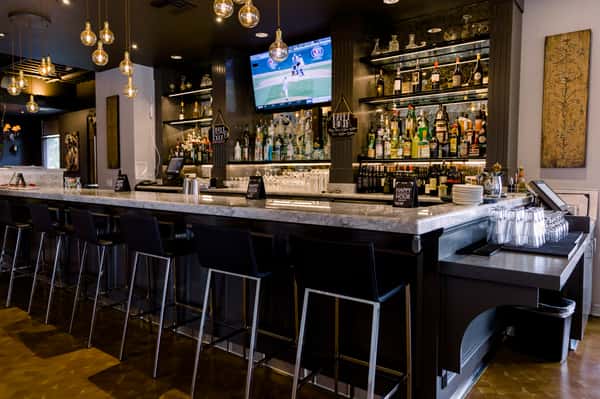 Modern bar interior with marble countertop, black barstools, hanging Edison bulbs, and fully stocked liquor shelves with TV above