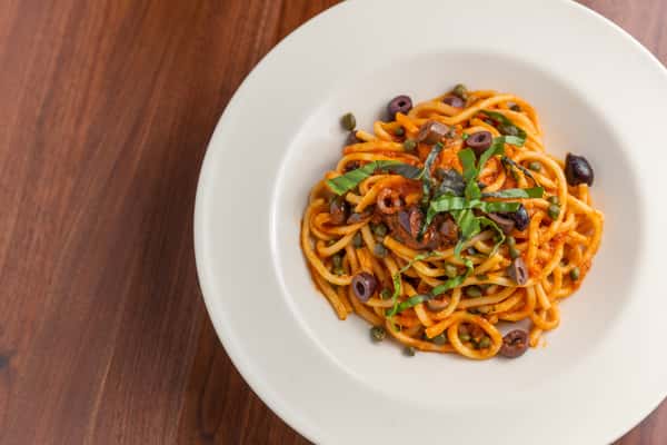 Spaghetti with tomato sauce, olives, capers, and fresh basil in white bowl on wooden table