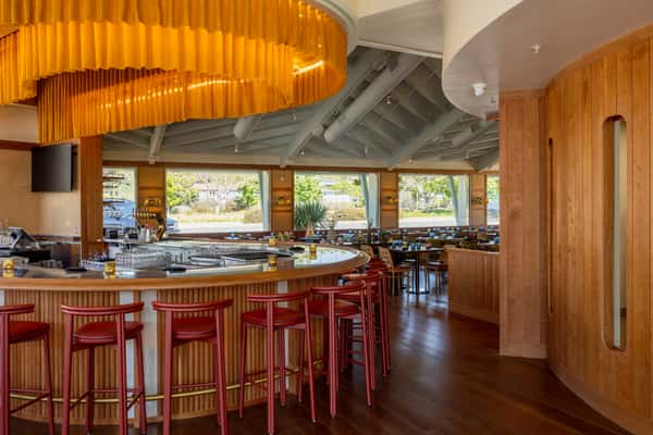 A modern restaurant interior featuring a circular bar, red stools, wooden accents, and large windows allowing natural light to illuminate the space.
