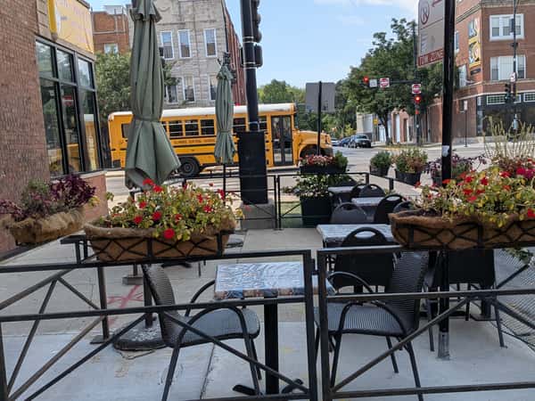 Sidewalk cafe seating area with tables, chairs, umbrellas, and flower planters, with a yellow school bus passing by in the background.