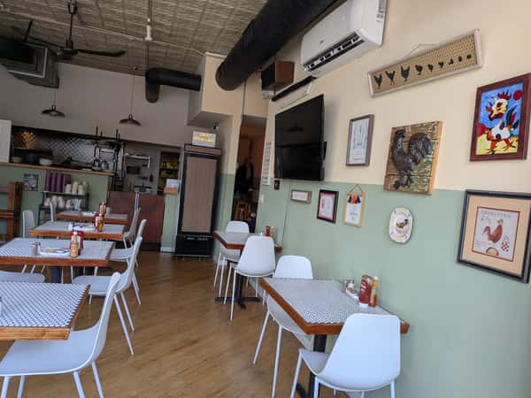 Cozy cafe interior with white chairs, patterned tables, rooster-themed wall art, and view into the open kitchen area.