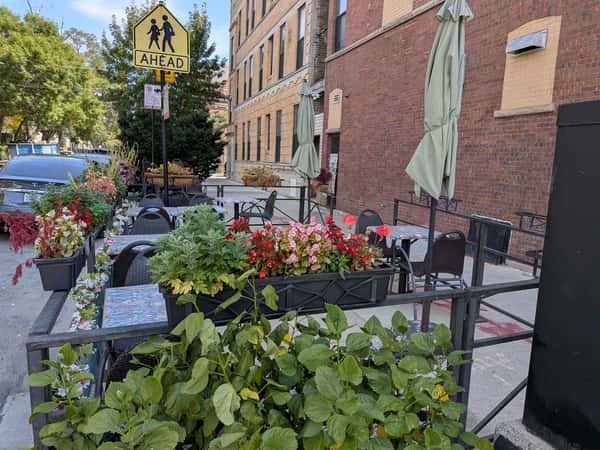 Outdoor cafe seating area with tables, umbrellas, and colorful flower planters along a city sidewalk.
