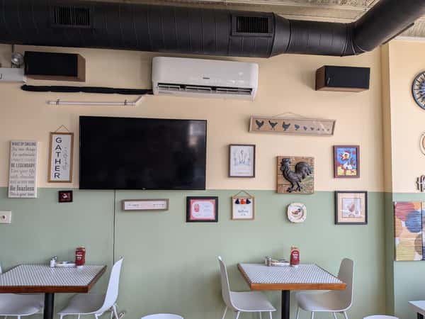 Cafe interior with white chairs, patterned tables, a wall-mounted TV, and rooster-themed artwork on the walls.
