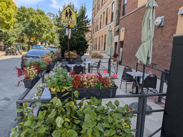 Outdoor cafe seating along a sidewalk with flower planters, closed umbrellas, and tables next to a brick building.