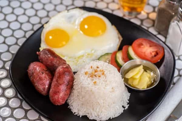 Breakfast plate with garlic rice, sunny-side-up eggs, sausages, sliced tomato and cucumber, and a small side of pickled vegetables.