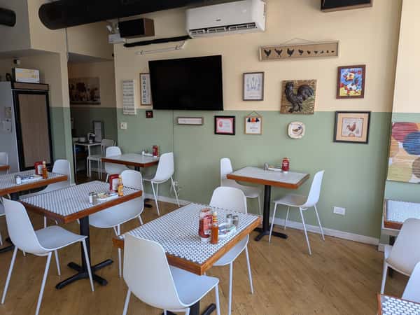 Cozy cafe interior with white chairs, patterned tables, wall art featuring roosters, and a mounted TV.