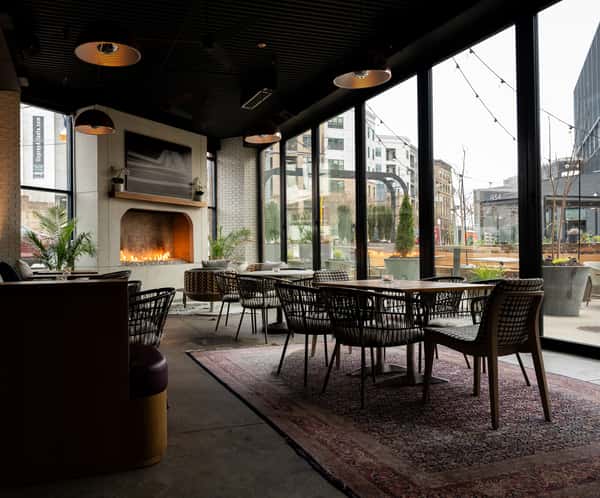 A cozy restaurant interior featuring a modern fireplace, tables with woven chairs, and large windows offering a view of the outside.