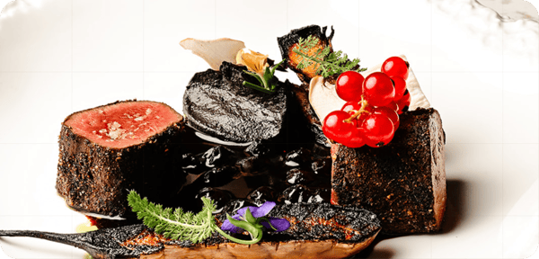 A close-up of an elegantly plated dish with seared meat cubes, blackened vegetables, garnished with herbs and red berries.