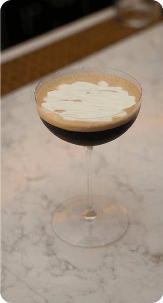 A close-up view of an espresso martini with a creamy froth on top, served in a coupe glass on a marble surface.