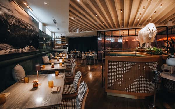 A modern restaurant with cozy seating, featuring tables with amber-colored candles, a large mountain photograph, and a mounted goat statue.