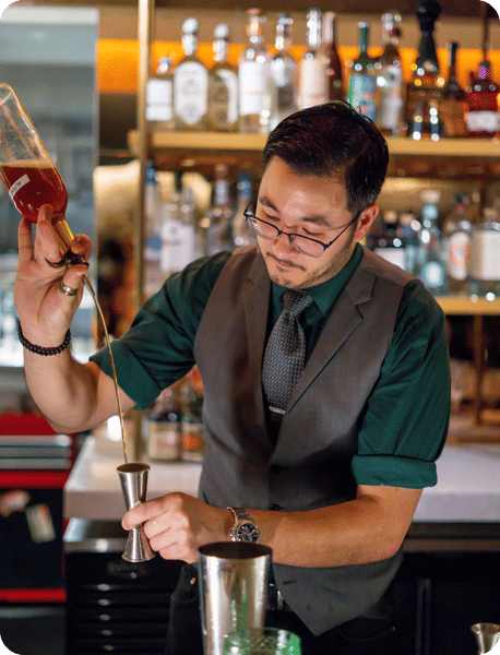 A bartender in a vest is carefully pouring a drink from a glass container into a jigger, with bottles of liquor in the background.