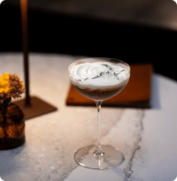 A close-up of a cocktail in a coupe glass with a creamy top and a light sprinkle of green garnish, sitting on a marble table with a small vase of flowers and soft lighting.