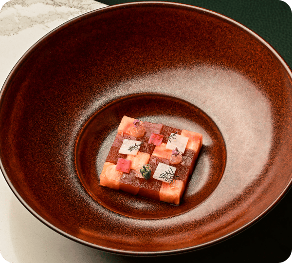 This dish appears to be a visually appealing arrangement of finely cut pieces of fish, likely sashimi, carefully arranged in a grid pattern within a large, deep, brown bowl. The top of the dish is garnished with delicate herbs and small flower petals, offering a vibrant contrast against the rich colors of the fish.
