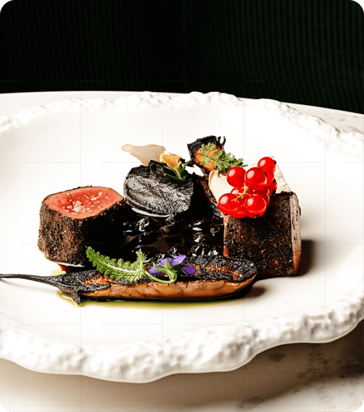 A close-up of an elegantly plated dish with seared meat cubes, blackened vegetables, garnished with herbs and red berries.