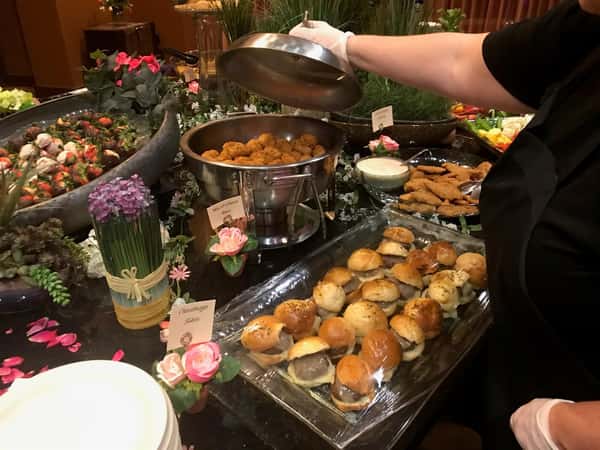 table of hot foods at an event including hamburger sliders and mac and cheese bites