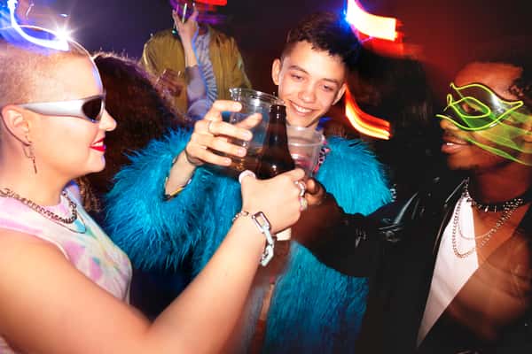 Three people toast drinks in a dark, neon-lit club, one in a fuzzy blue coat, one in reflective sunglasses, and one in an LED mask.