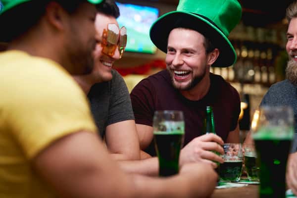 Three men at a bar, laughing and drinking green beer. One wears a green hat, another wears beer mug novelty glasses.