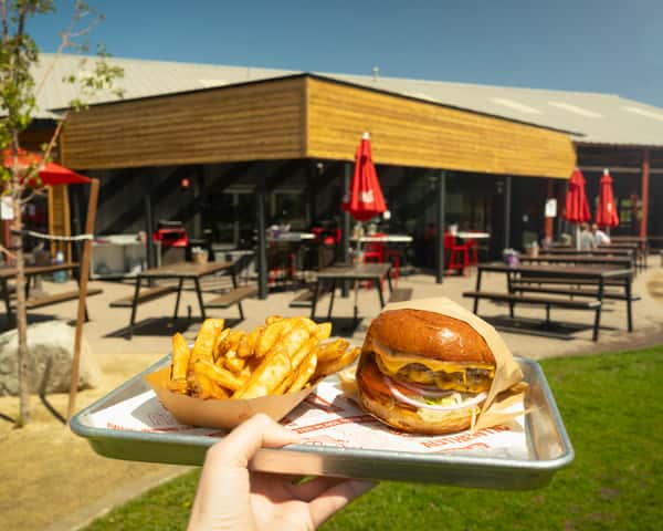 hand holding a double roadside cheeseburger and a side of fries with the patio in the background.