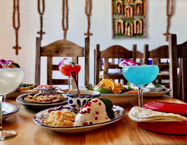 Traditional Mexican table setting with enchiladas, rice, tortillas, margaritas, and rustic wooden chairs in dining room.