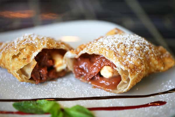 Golden fried Mexican empanadas filled with sweet fruit and cream, dusted with powdered sugar and plated with chocolate drizzle.