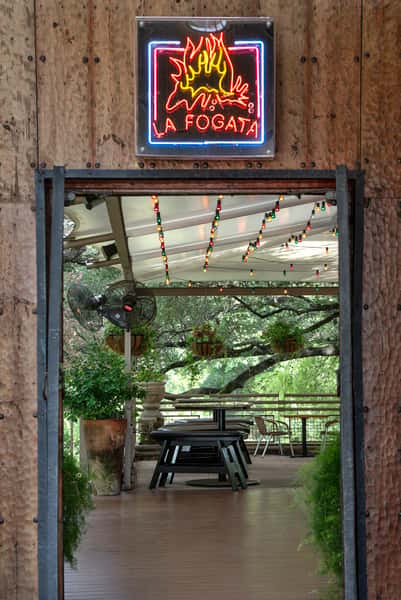 Neon La Fogata sign above rustic entrance leading into a tree-shaded outdoor Mexican restaurant patio with string lights.