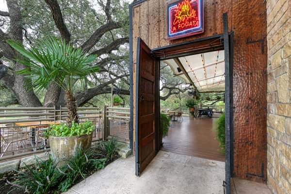 Open-air Mexican restaurant patio entrance with rustic wooden doors, glowing La Fogata neon sign, string lights, and tree-shaded outdoor seating surrounded by greenery.