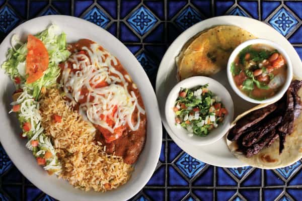 Authentic Mexican food plates featuring a cheese-topped enchilada with red sauce, Mexican rice, refried beans, fresh lettuce and tomato, plus a steak taco with pico de gallo and charro beans.