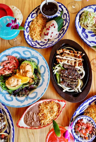 Colorful Mexican restaurant spread with grilled steak fajitas, chicken enchilada with white sauce, Mexican rice, refried beans, fresh salad, tacos, and vibrant margaritas on a wooden table