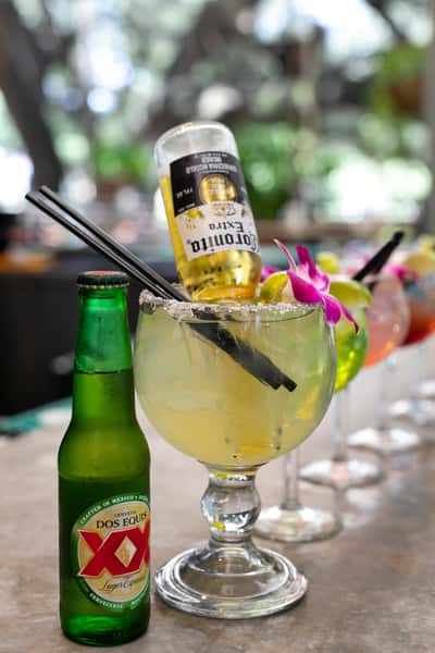 Classic Mexican margarita topped with an upside-down Corona Extra bottle beside a Dos Equis beer on an outdoor patio table.
