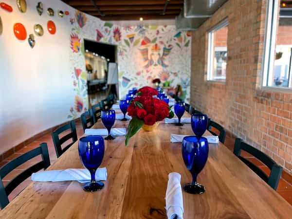 Private dining table set with blue glassware, red floral centerpiece, and hand-painted Mexican mural backdrop.