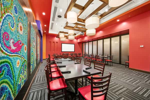 Modern private dining room in a Mexican restaurant with long tables, red accent walls, colorful mosaic art panels, wood ceiling beams, and contemporary pendant lighting.