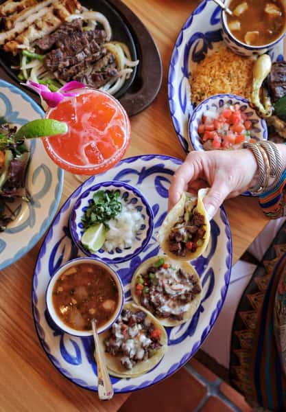 Hand holding a carne asada taco over a Mexican table spread with tacos, caldo soup, pico de gallo, rice, and a bright red margarita.