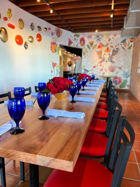 Long communal dining table set with blue glassware, red cushioned chairs, folded napkins, and rose centerpieces inside a colorful Mexican restaurant private dining space with mural walls.