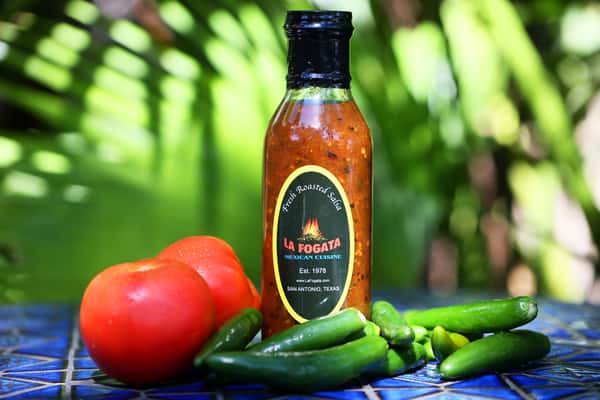 Bottle of La Fogata fresh roasted salsa surrounded by ripe tomatoes and jalapeño peppers on a tiled table.